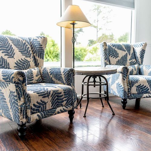 Two patterned armchairs facing a small table with a lamp; a bright window and wooden floor complete the cozy scene.