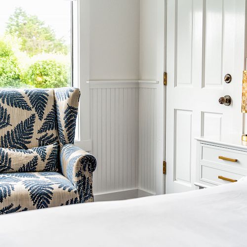 The image shows a cozy bedroom corner with a patterned armchair, a nightstand with a lamp, and a door leading outside, all in a bright and airy setting.