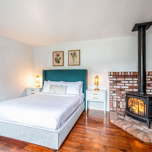 A cozy bedroom with a bed, nightstands, framed art on the walls, and a small wood-burning stove set against a brick wall in the corner.