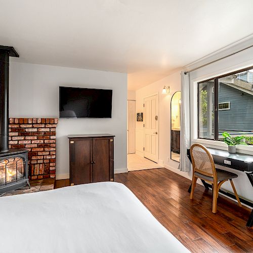 This image shows a cozy bedroom with a wood-burning stove, TV, wooden desk and chair by a window, and a white bed, all on a hardwood floor.