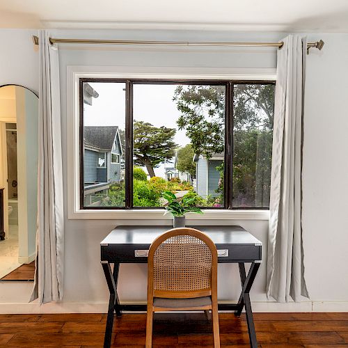 A room with a window, desk, chair, and a large mirror on the left wall. Outside, trees and houses are visible through the window.