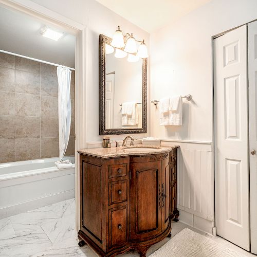 A bathroom features a wooden vanity with a mirror, dual-light fixture, towels, and a tub with a curtain in a tiled shower area.