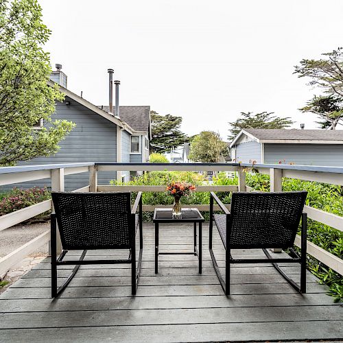 A wooden deck with two black chairs and a small table overlooking a garden, houses, and distant trees under a cloudy sky.