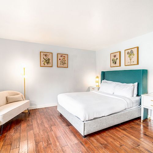 A bright bedroom with a bed, green headboard, two nightstands, a chair, three botanical wall art pieces, and wooden flooring.