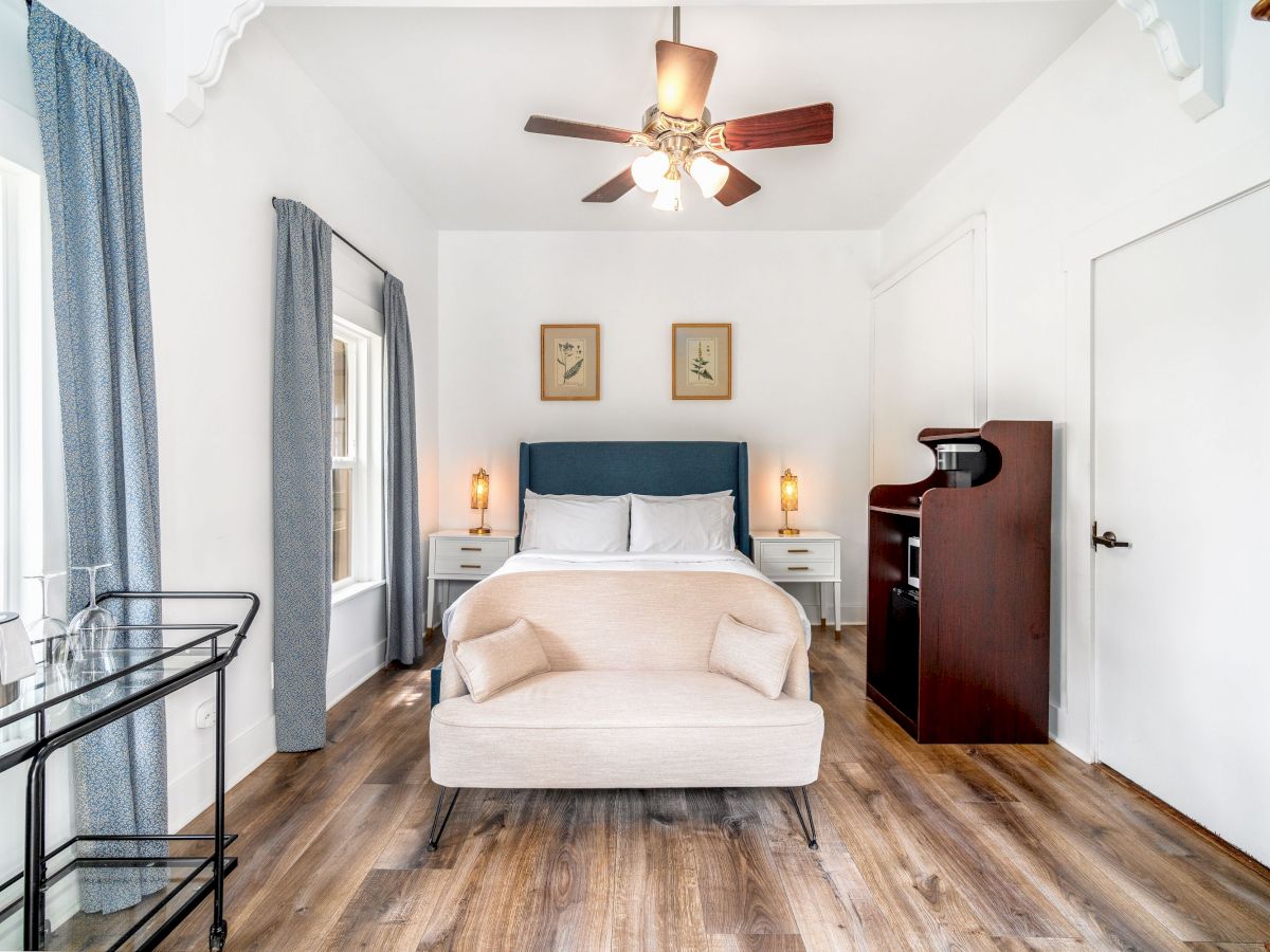 A bright bedroom with a double bed, bedside tables, sofa, ceiling fan, framed pictures, and a shelf unit. The floor is wooden, and windows are curtained.