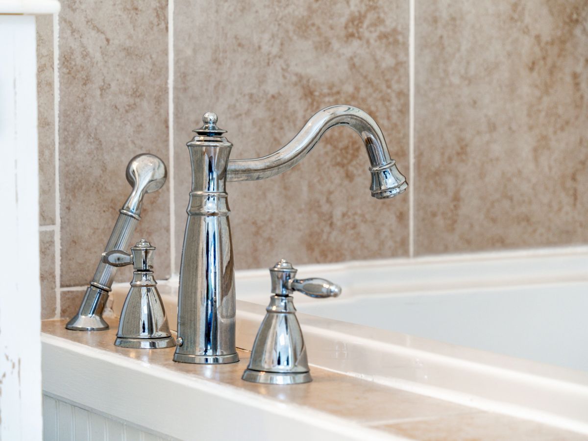 The image shows a close-up of a bathroom faucet set, including a spout and handles, with a bathtub and tiled wall in the background.