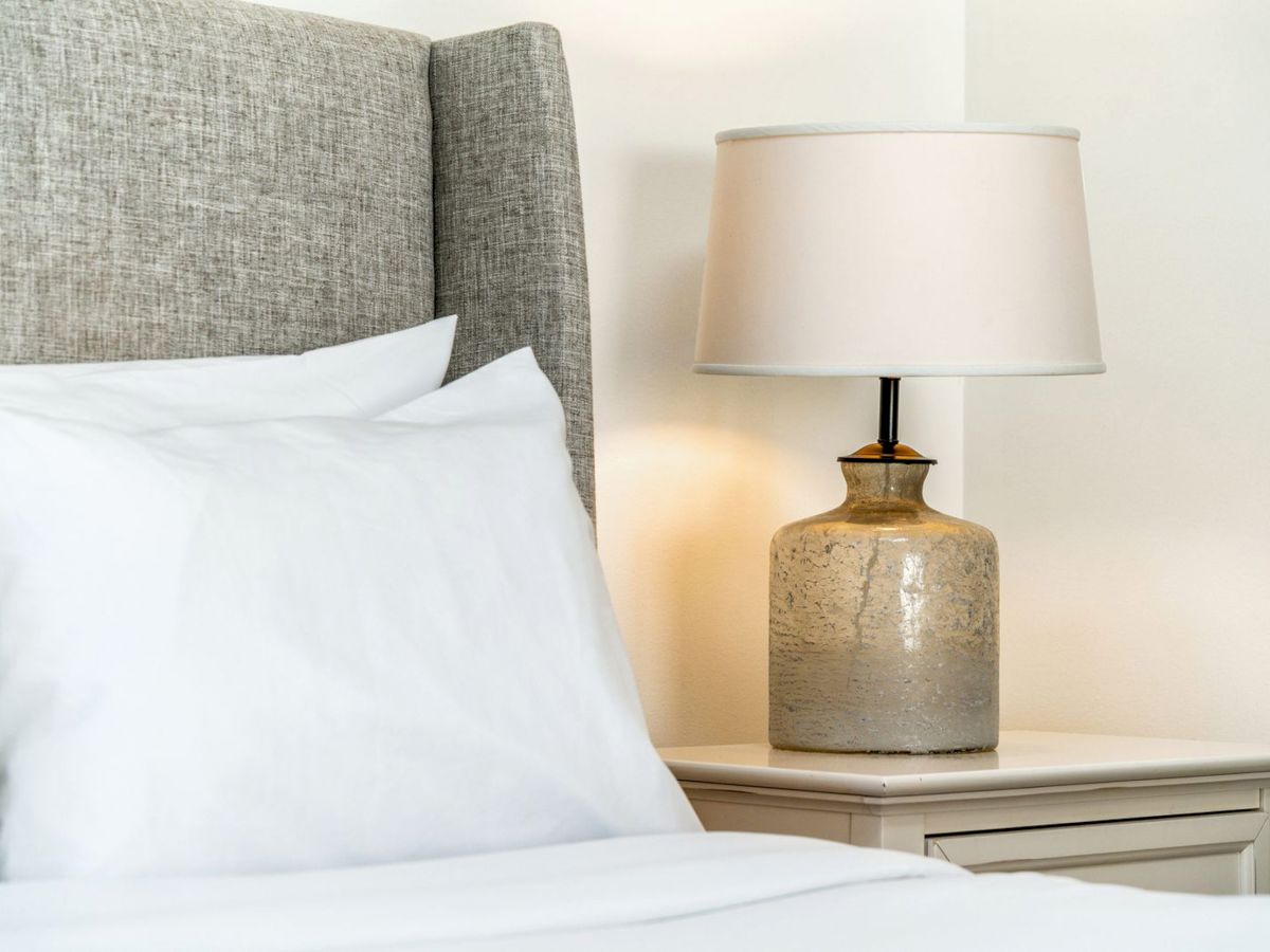 A neatly made bed with white linens, a grey upholstered headboard, and a ceramic lamp on a bedside table.