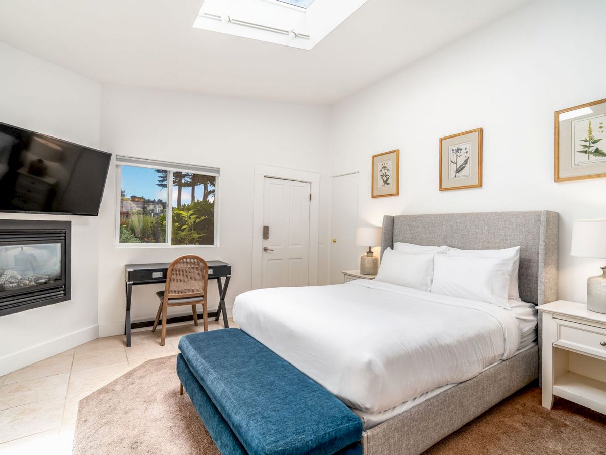 A bright, modern bedroom with a skylight, large bed, wall-mounted TV, fireplace, desk and chair by a window, and two nightstands with lamps.