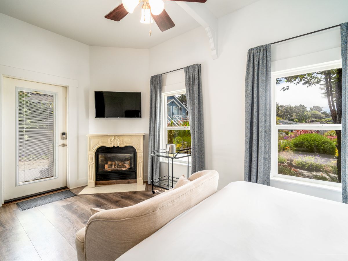 A cozy bedroom features a bed, a fireplace, a mounted TV, large windows with curtains, a door, and a ceiling fan, with garden views outside.