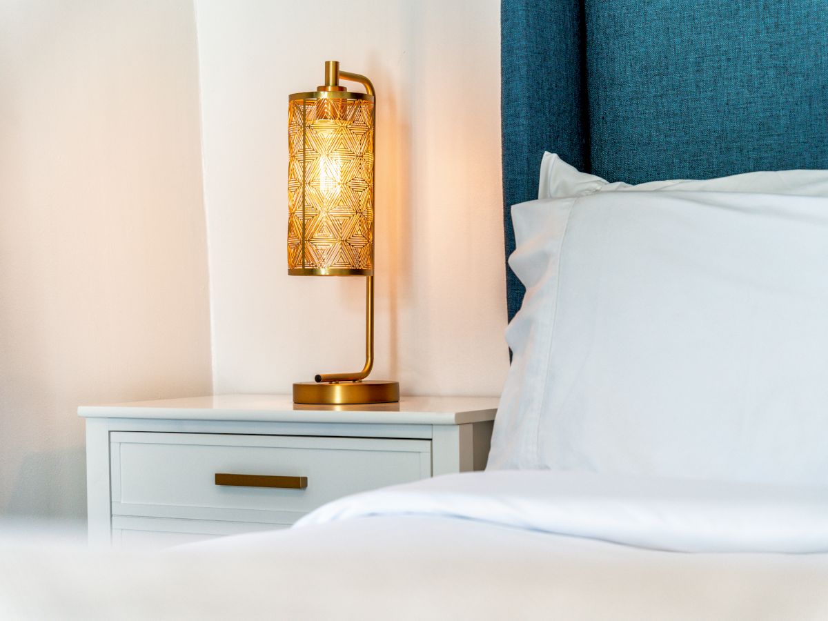 A bedside table with a white drawer, a decorative lamp, and a bed with white sheets and a blue headboard are visible in the image.