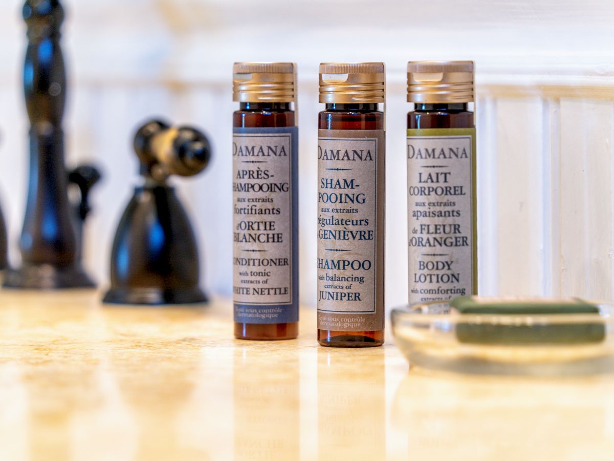 Three small bottles of Damana toiletries: conditioner, shampoo, and body lotion, are placed on a bathroom countertop next to a bar of soap.