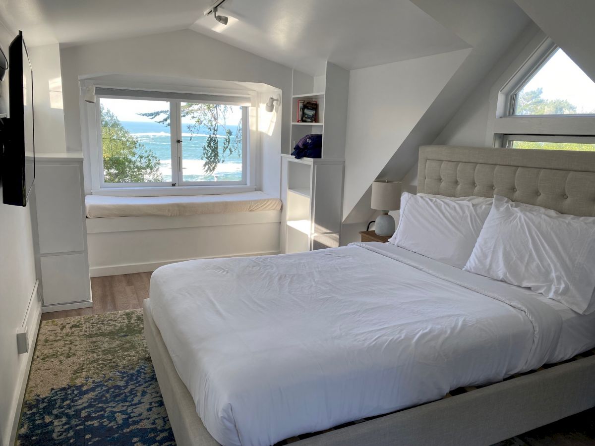 A cozy bedroom with a white bed, windows offering a scenic view of water, and shelves on either side. There's a bench seat under the window.