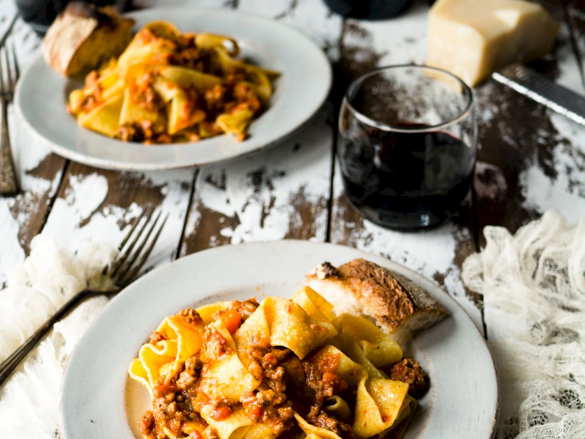 Two plates of pasta with bread, two glasses of red wine, a fork, cheese, and a rustic table setting complete the scene.