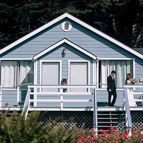 A blue and white house with two people standing on the front porch, surrounded by greenery and vibrant flowers.