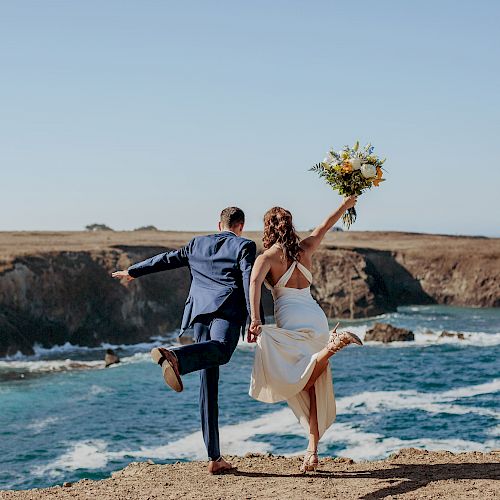 A couple, dressed formally, stands with one leg up, facing the ocean on a rocky cliff, while the woman holds a bouquet of flowers.