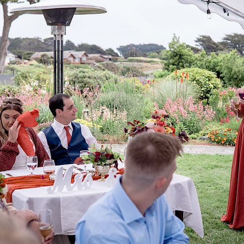 A woman in a red dress is giving a speech at an outdoor event. Several people are seated and listening attentively at decorated tables.