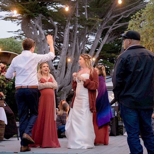 People are attending an outdoor event with string lights overhead. Some are dancing, while others are standing and talking.