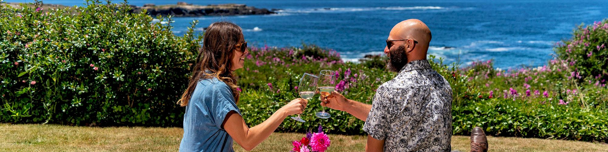 Two people having a picnic on a blanket by the sea, clinking glasses with a scenic ocean view and clear blue sky.