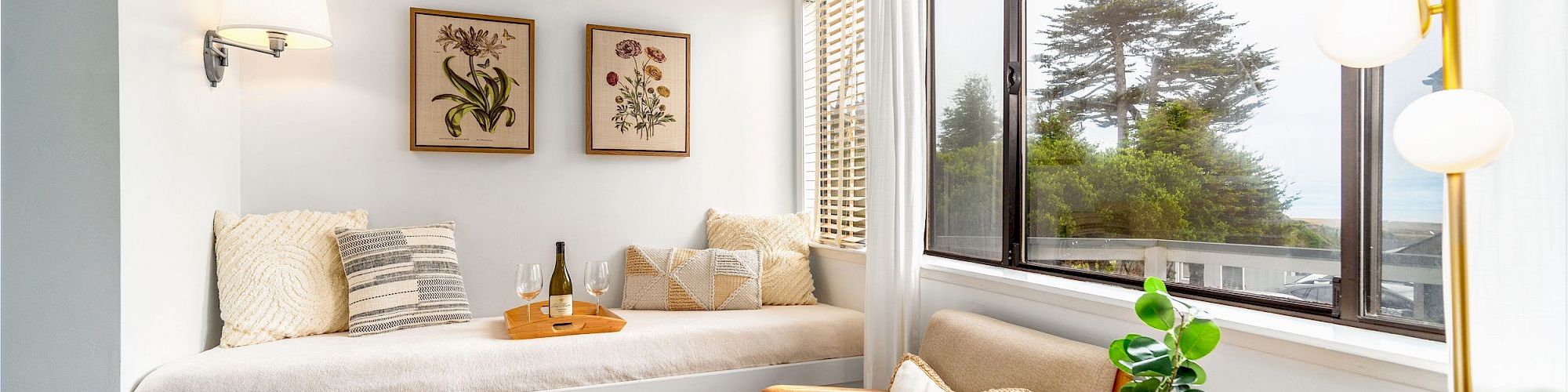 A cozy sitting nook with a daybed, pillows, wall art, window views, a plant, a chair, and a lamp on a small table, creating a serene atmosphere.