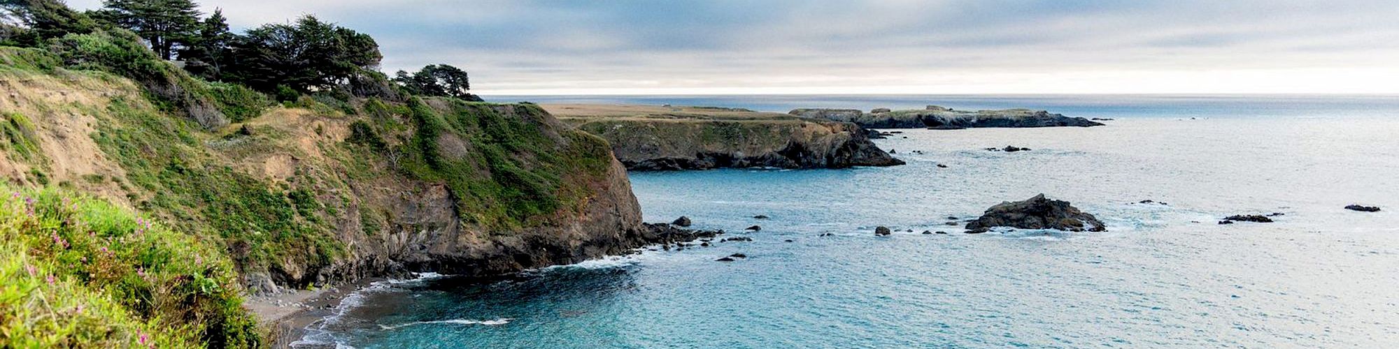 A scenic coastal landscape featuring rugged cliffs overlooking a serene, blue ocean under a partly cloudy sky can be seen in the image.