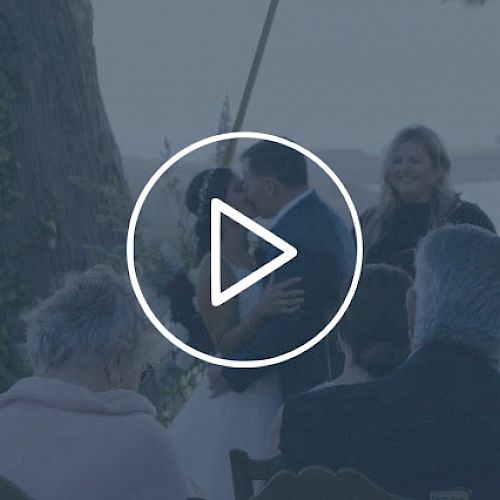 A couple is getting married outdoors, with a few guests watching. A play button is overlaid, suggesting this is a video screenshot.