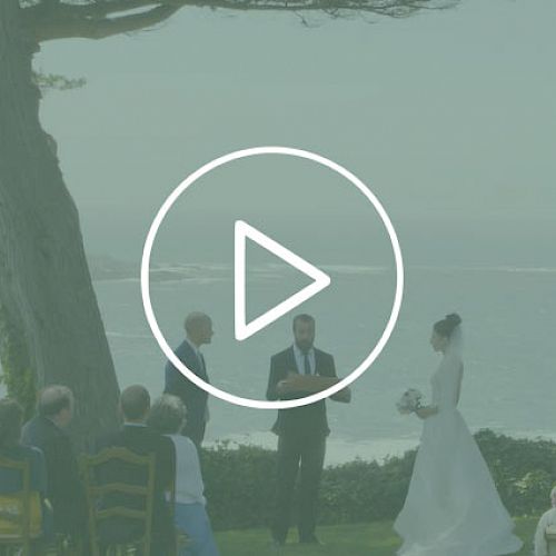 A wedding ceremony taking place outdoors with the ocean in the background. The couple stands before an officiant, and guests are seated nearby.