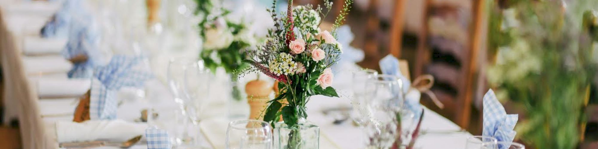 A long banquet table is elegantly set with glassware, floral centerpieces, blue checkered napkins, and wooden chairs, ready for a gathering.