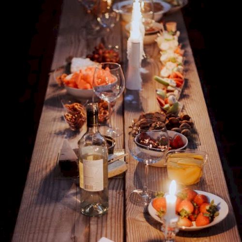 A long wooden table is set with various dishes, candles, and a bottle of wine for a cozy dinner setting.