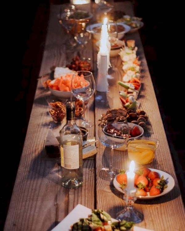 A long wooden table is set with various dishes, candles, and a bottle of wine for a cozy dinner setting.