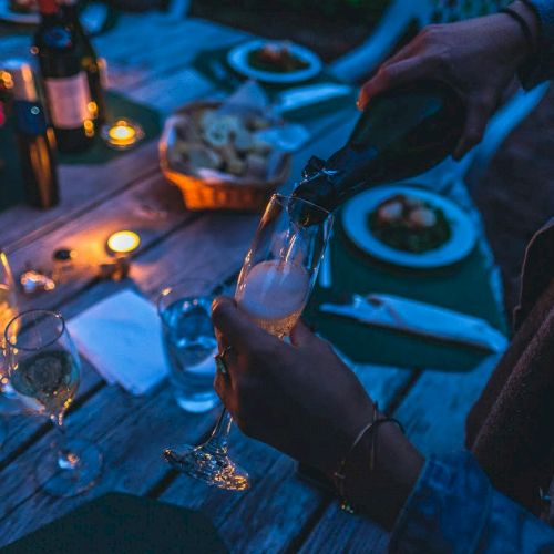 The image shows a dinner table setting with candles, wine bottles, dishes, and someone pouring champagne into a flute glass.