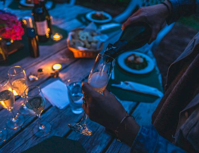 The image shows a dinner table setting with candles, wine bottles, dishes, and someone pouring champagne into a flute glass.