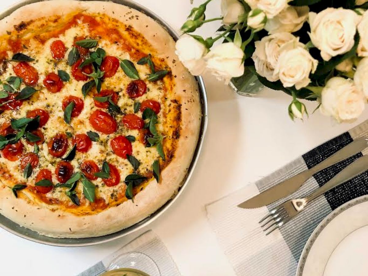 A pizza with tomatoes and basil, a glass of white wine, a bouquet of white roses, and a neatly set table with plates and cutlery.