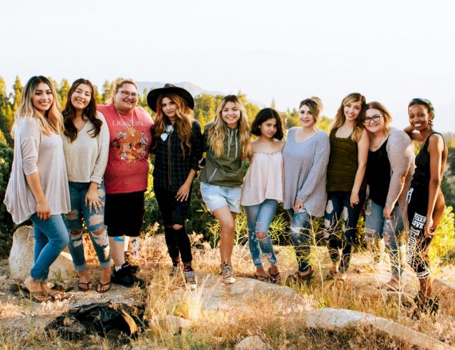 A group of people smiling outdoors, standing on rocky terrain with greenery in the background.