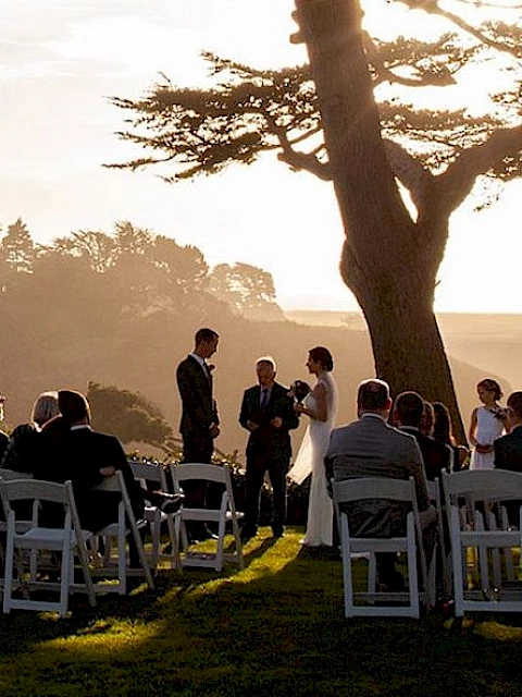 A group of people seated outdoors, appearing to attend a ceremony under large trees with a scenic sunset view in the background.