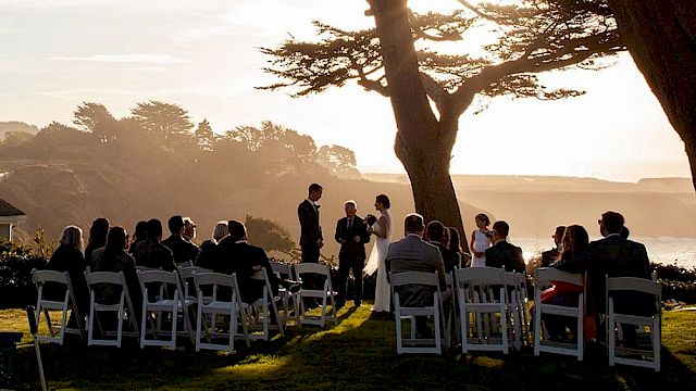 A group of people seated outdoors, appearing to attend a ceremony under large trees with a scenic sunset view in the background.