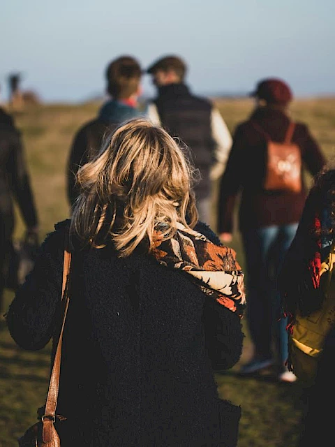 A group of people walking outdoors on a grassy area, with backpacks and jackets, in a casual, relaxed setting.