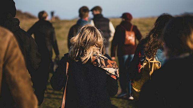 A group of people walking outdoors on a grassy area, with backpacks and jackets, in a casual, relaxed setting.