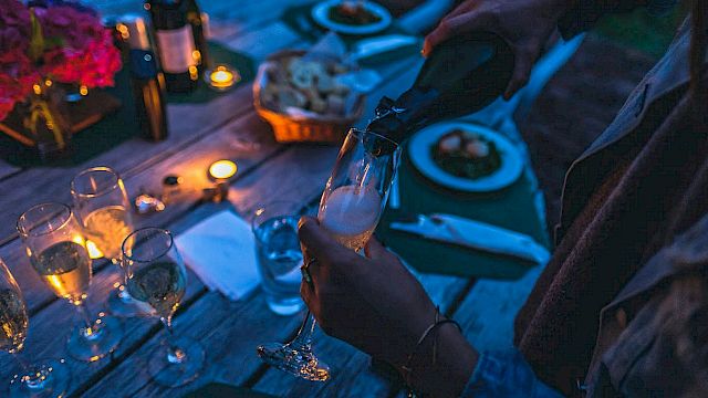 A person pours champagne at a dimly lit outdoor dinner table with candles, wine bottles, flowers, and plates of food.