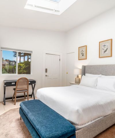 A bedroom with a bed, nightstands, desk, TV, and fireplace. Decor features a skylight, framed art, and a window with a view of palm trees.