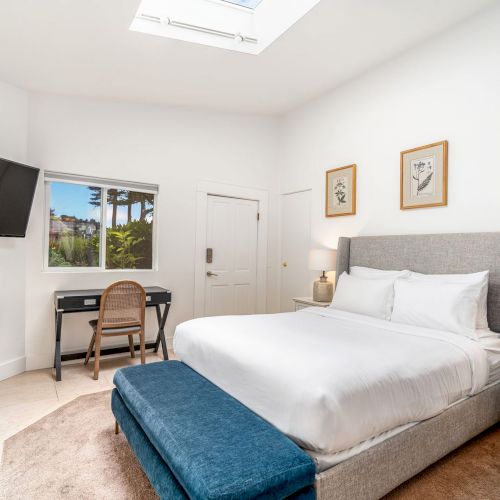 A bedroom with a bed, nightstands, desk, TV, and fireplace. Decor features a skylight, framed art, and a window with a view of palm trees.