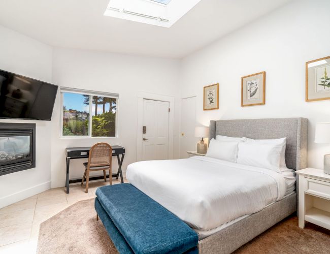 A bedroom with a bed, nightstands, desk, TV, and fireplace. Decor features a skylight, framed art, and a window with a view of palm trees.