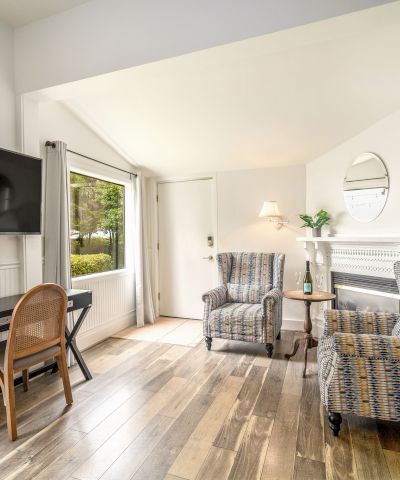 The image shows a bright room with two armchairs, a wall-mounted TV, a desk, and a fireplace, featuring wooden flooring and decor.