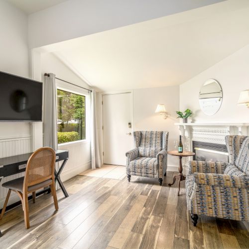 The image shows a bright room with two armchairs, a wall-mounted TV, a desk, and a fireplace, featuring wooden flooring and decor.