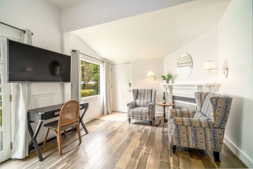 The image shows a bright room with two armchairs, a wall-mounted TV, a desk, and a fireplace, featuring wooden flooring and decor.