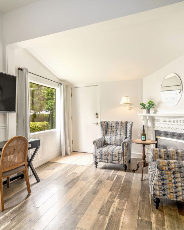 The image shows a bright room with two armchairs, a wall-mounted TV, a desk, and a fireplace, featuring wooden flooring and decor.