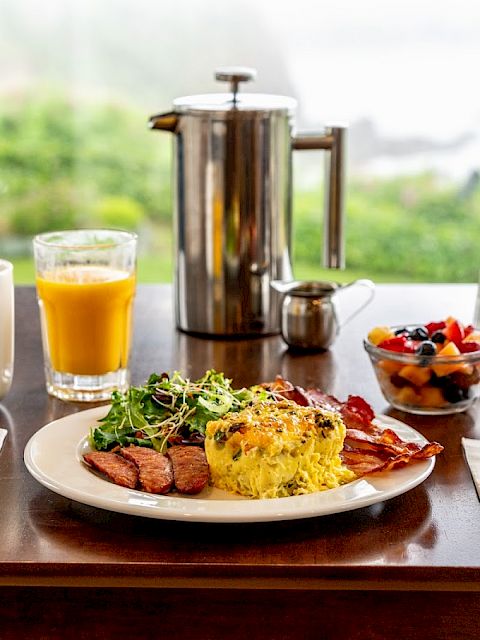 A breakfast with eggs, bacon, sausage, salad, and fruit served with coffee and orange juice on a wooden table by a scenic view.