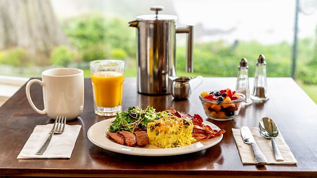 A breakfast with eggs, bacon, sausage, salad, and fruit served with coffee and orange juice on a wooden table by a scenic view.