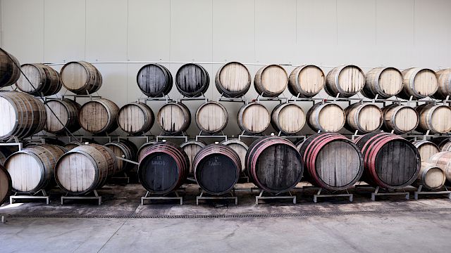 The image shows rows of stacked wooden barrels in a storage area, likely for wine or spirits aging, set against a plain wall.