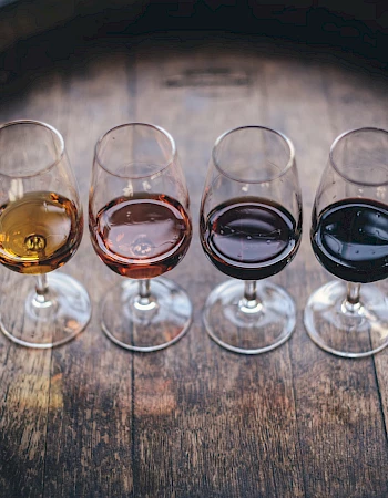 Four wine glasses with varying shades of wine sit on a wooden barrel surface.