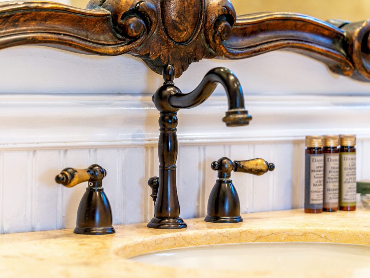 A vintage bathroom sink with a dark metal faucet and handles, a light marble counter, beadboard wall, and small bottles on the right, classy and rustic.